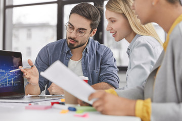 Cropped shot of successful female and male enterpreneurs study company`s budget, focused on screen of laptop as analyze graphic together, work with business papers, sit against office interior