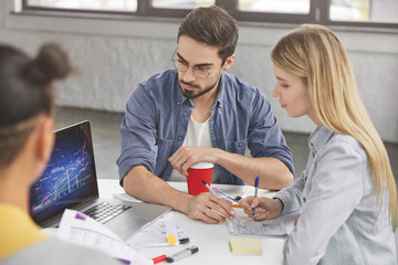 Skilled young marketing experts analyze information together, cooperate to prepare business project, create graphics to illustrate financilal situation. Mixed race team members speak at working desk