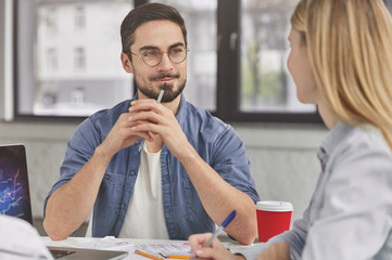 Attentive pleased brunet male listens attentively to female banker who explains bank facilities. Female and male coworkers talk about sales figures, work with modern technologies and papers.