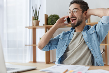 Bearded positive male manager talks with close friend on smart phone, has break after work, checks...