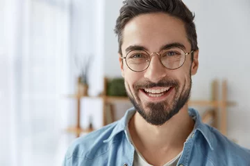 Fotobehang Close up shot of cheerful satisfied attractive male with stubble, has broad smile, wears round spectacles, rejoices success at work, stands against cozy interior. Fashionable designer glad be praised © Wayhome Studio
