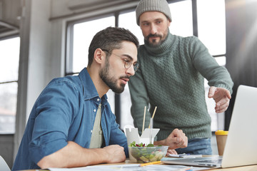 Photo of male colleagues generate innovative ideas for creating new startup, collaborate during working process, have snack. Unshaven stylish male demonstrates his work to business partner, ask advice