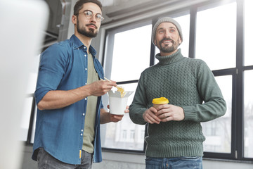 Two men coworkers stand in spacious office lobby, eat junk food and drink hot coffee, look into distance as observe presentation. Male colleagues during working break have business conversation