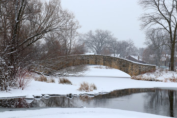 Scenic winter view in a city park. Winter snowy day landscape with an old style brick bridge during snowfall. Tenney Park in the city of Madison, the capital of Wisconsin, Midwest USA.