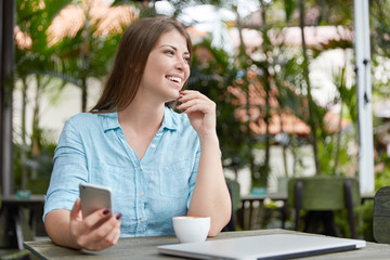 Adorable female model looks happily aside, checks newsfeed on modern mobile phone, sits in outdoor cafeteria, enjoys vacations, uses free wifi. Glad woman posts new photos on webpage, shares news