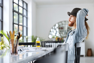 Horizontal shot of fashionable male model in elegant hat sits at table, looks pensively in window, enjoys weekends, thinks about creating presentation, has specific appearance and long hair.