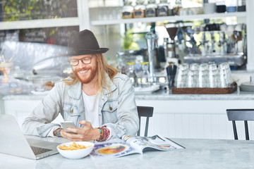 Talented male developer has ginger beard and mustache, dressed in fashionable clothes, has lunch at cafe, eats fast food, works on laptop computer, recieves message from friend on cell phone