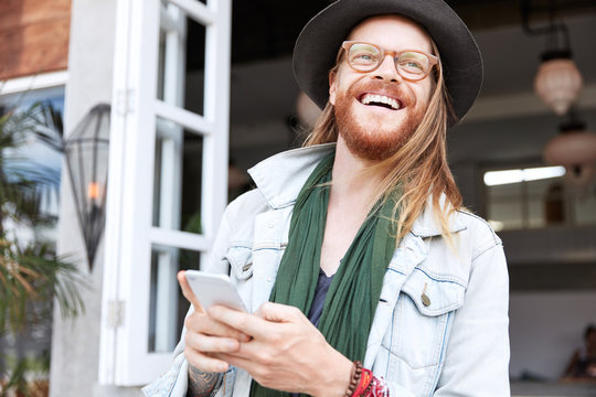 Fashionable hipster guy dressed in stylish black hat, denim shirt, spectacles, has long hair and ginger thick bearded, mustache, looks happpily aside, recieves text message with good news on cellular