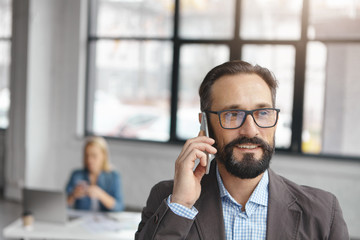 Close up portrait of bearded mature businessman has telephone conversation with partner, discuss future transactions or net capital gain, stands against office interior, people work in background