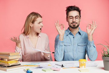 Angry irritated pretty female student looks at male partner who meditates at working table, tries...