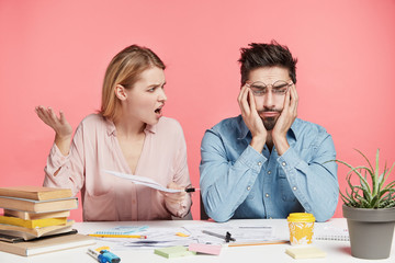 Frustrated puzzled male has problems with studying, feels badly and his female groupmate shouts at him for bad work, being dissatisfied with prepared project, isolated over pink studio background