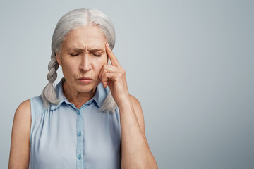 Concentrated forgetful grey haired mature woman with two plaits, keeps finger on temple, closes...
