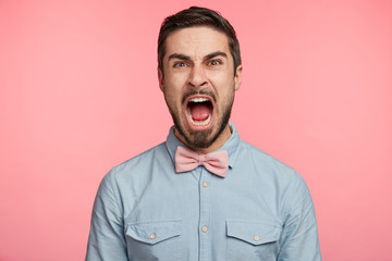 Obraz premium Annoyed brunet male yells with anger as being dissatisfied with working conditions, dressed formally, poses against pink background. Discontent mad man screams loudly at someone. Negative emotions