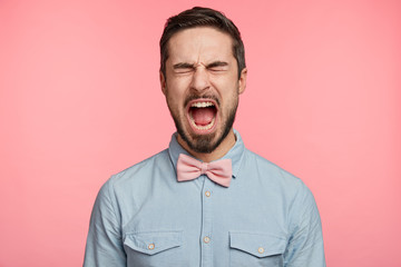 Emotional dissatisfied male with thick dark beard, closes eyes and keeps jaw dropped out, expresses his great dissatisfaction with something, dressed in formal shirt and bowtie poses against pink wall