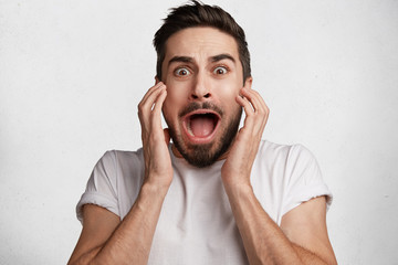 Emotional scared brunet male has dark stubble, looks with frightened expression as sees something...