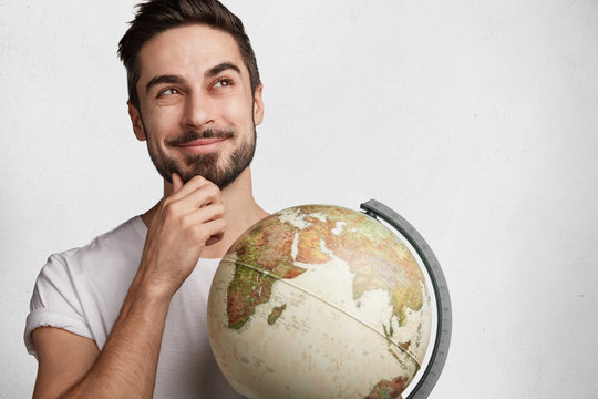 Dreamy hipster guy with globe thinks about which new country to visit, being in good mood as has vacations, isolated over white background. Shot of handsome male tourist. World wide concept.