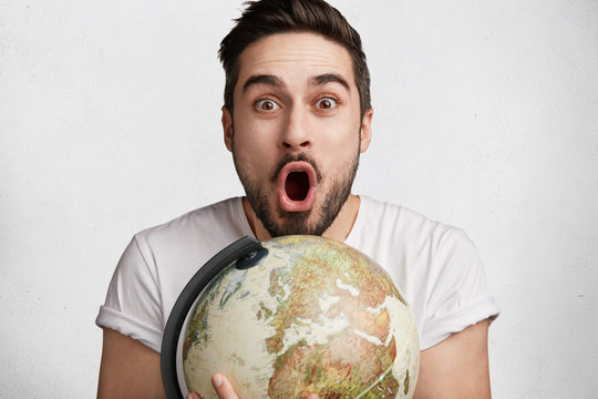 Shocked brunet male embraces globe, being surprised to win tourist voucher in exotic country, isolated over white background. Surprised young geography teacher poses indoor. Wordwide concept