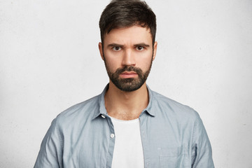 Studio shot of serious strict male with dark beard and mustache, looks directly into camera, listens seriously interlocutor, disagrees with somebody`s opinion, poses against white concerte wall