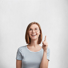 Pleased smiling woman with bobbed hairdo, dressed casually, happy to make repairment in her room, shows at new ceiling, being in good mood, isolated over white concrete wall, copy space for your text