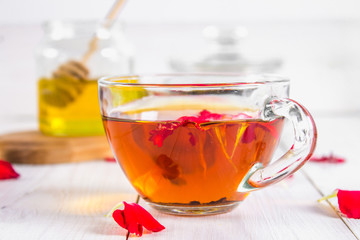 A cup of tea, in the background of a bank of honey and a jar with a black herbal floral tea on a white wooden table.