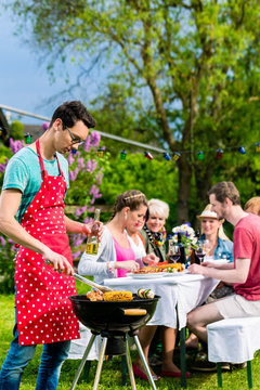 Man grilling meat on garden barbecue party, in the background friends eating and drinking