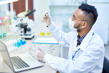 Side view portrait of young Middle-Eastern scientist working with test tubes and laptop studying...