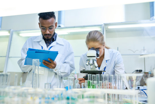 Portrait of young Middle-eastern scientist taking notes on clipboard while working on medical research in laboratory while his female colleague looking in microscope