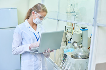 Portrait of blonde female scientist using laptop while working in medical laboratory doing internet research, copy space