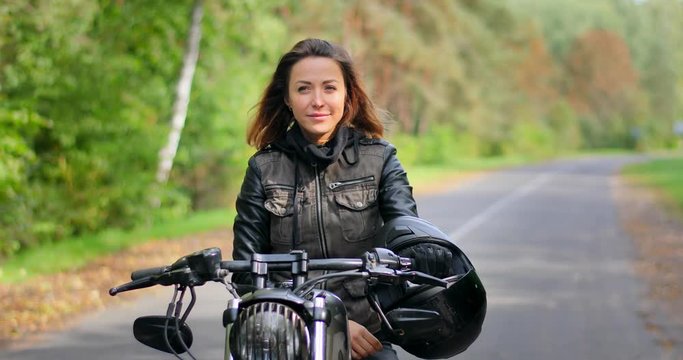 4k Woman sits on cafe racer custom motorbike smiling touching hair autumn nature forest road. Girl biker on motorcycle posing starts engine hands on handlebars ready to ride sunny defocused background