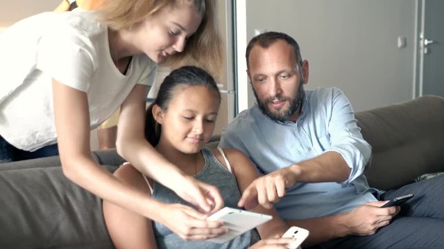 Father with his daughters talking, using tablet and smartphone on sofa at home
