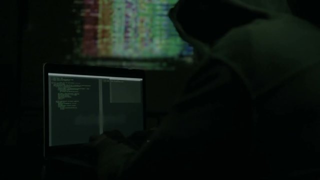 Male hacker working on a computer while green code characters reflect on his face in a dark office room.
