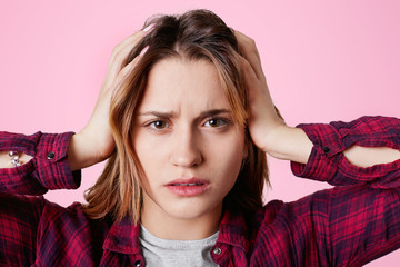 Fototapeta premium Close up portrait of stressful female model touches head and suffers from headache, looks frusrtated at camera, dressed in checkered shirt, isolated over pink background. Emotional woman indoor