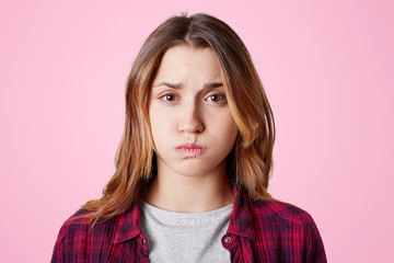 Studio shot of pretty young female blows cheeks, looks with discontent expression, being offended or angry with someone, dressed in casual clothes, isolated over studio pink background.