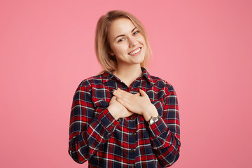 Romantic beautiful young female with pleasant shining smile, keeps hands on chest, dressed casually, demonstrates her friendly feeling to people who surround her, isolated over pink background