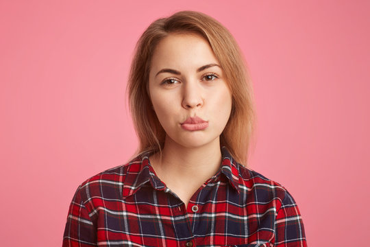 Headshot of abused female with short hair, curves lips with dissatisfaction, being offended with her boyfriend who didn`t come on date, dressed in casual checkerd shirt, isolated on pink wall