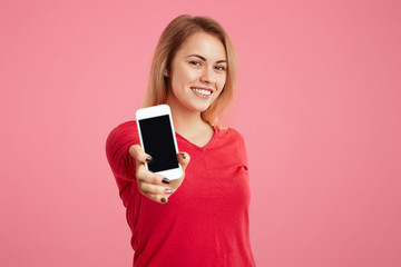 Pretty female with shining smile shows smart phone with blank copy screen for your advertisment, glad with its functions, isolated over pink background. People, technology, advertising concept