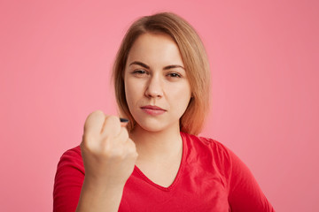 Serious angry woman puts up fist as tries to warn you, dressed in red sweater, isolated over pink background. Pleasant looking female shows fist, being strict and aggressive, tries to show her power