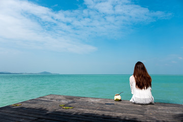 A beautiful asian woman on white dress sitting and looking at the sea and blue sky on wooden balcony with feeling relaxed