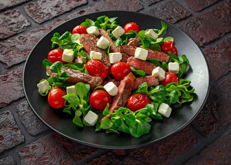 Juicy Beef Sirloin Steak Salad with roasted tomatoes, feta cheese and green vegetables in a black plate. healthy food