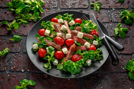 Juicy Beef Sirloin Steak Salad with roasted tomatoes, feta cheese and green vegetables in a black plate. healthy food