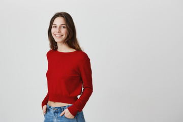 Close up of beautiful young attractive girl with dark long hair i red sweater and jeans smiling, holding hands in pockets, looking in camera with relaxed and happy expression