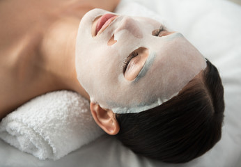 Close up of calm young woman lying with textile moisturizing mask at wellness center