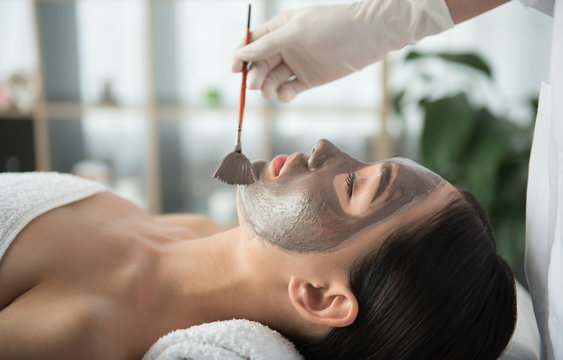 Side View Of Asian Girl Getting Skin Treatment At Beauty Salon. Beautician Is Holding Brush Near Her Face. Healthy Facial Mask Concept