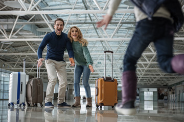 Low angle of joyous kid meeting lovely mother and father at the airport. Girl is running towards them