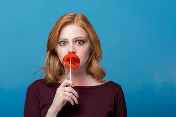 Fashion portrait young woman and lollipop is having fun over colorful blue background. Redhead girl...
