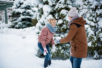Fototapeta na wymiar Happy young man and woman are having fun with snow in winter forest. They are looking at each other and smiling