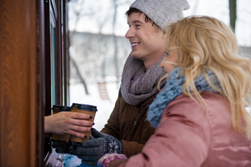 Side view of excited young man and woman are buying hot beverage in shop outside. They are taking cups and laughing. Winter date concept