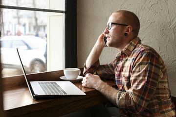 Fototapeta premium Smiling bearded businessman wearing casual hipster clothing using laptop and cell smartphone in coffe house.