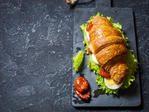 Croissant sandwich with tuna, hard boiled egg, salad and sun-dried tomatoes on stone table