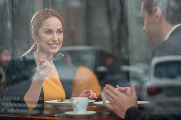 Portrait of cheerful lady eating appetizing meal while speaking with colleague. They sitting at table in cafe. Communication and leisure concept